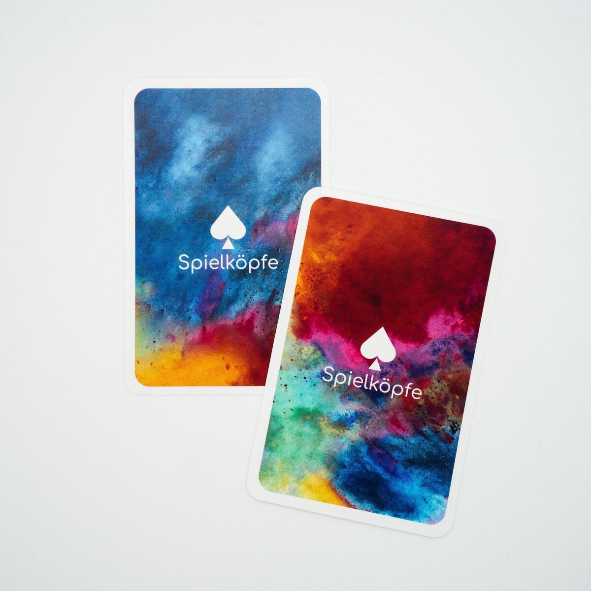 Zwei Spielkarten des Spiels Rommé zeigen bunte, abstrakte Kunst auf der Rückseite, mit 'Spielköpfe' und einem Pik-Symbol aufgedruckt.