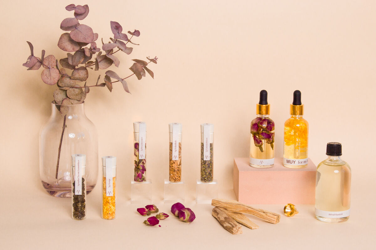 Ein Naturkosmetik-Set namens »MUY« auf einem minimalistischen Hintergrund. Verschiedene Glasflaschen und Reagenzgläser mit Kräutern und Blumenblättern stehen zusammen mit Eukalyptus-Zweigen in einer Vase. Enthält Seren, Öle und weitere natürliche Pflegeprodukte.