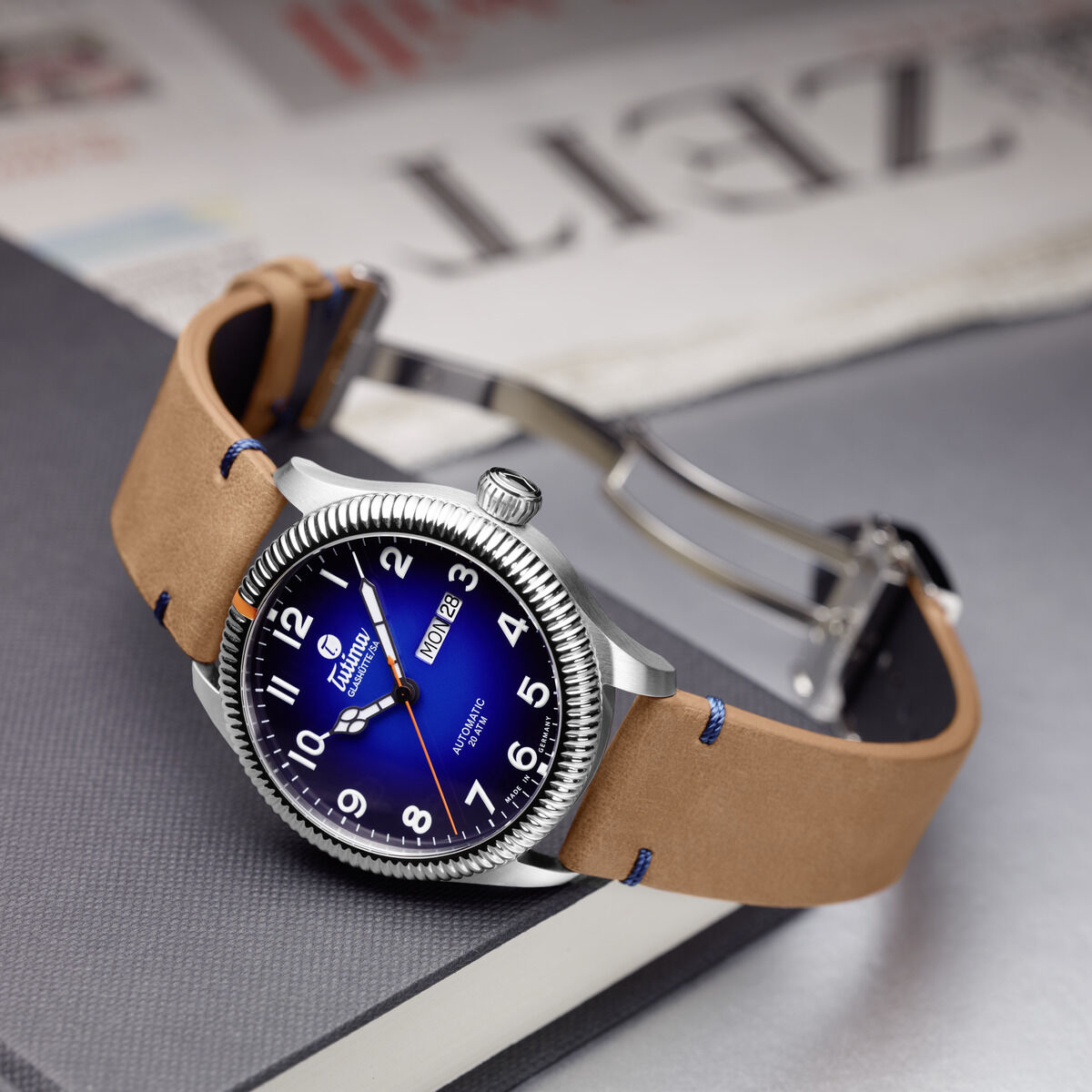 Die Tutima Glashütte ZEIT-Sonderedition Uhr 'Grand Flieger Classic Automatic' mit blauem Zifferblatt, silbernem Edelstahlgehäuse und hellbraunem Lederarmband liegt auf einem grauen Buch, im Hintergrund eine unscharfe Zeitung mit der Überschrift 'ZEIT'.