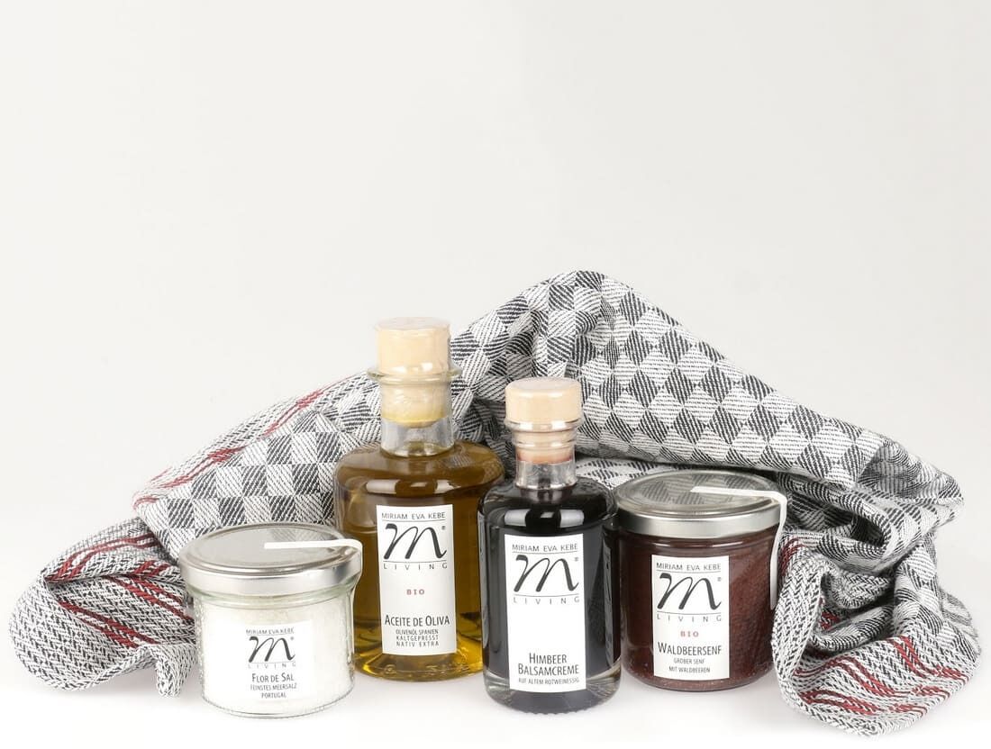 Das Jubiläums-Set »Zeit für fruchtige Würze« enthält eine Flasche mit Öl, eine Flasche mit Essig, ein Glas mit Fruchtaufstrich und ein Glas mit Salz, die in einem schwarz-weiß karierten Handtuch arrangiert sind.