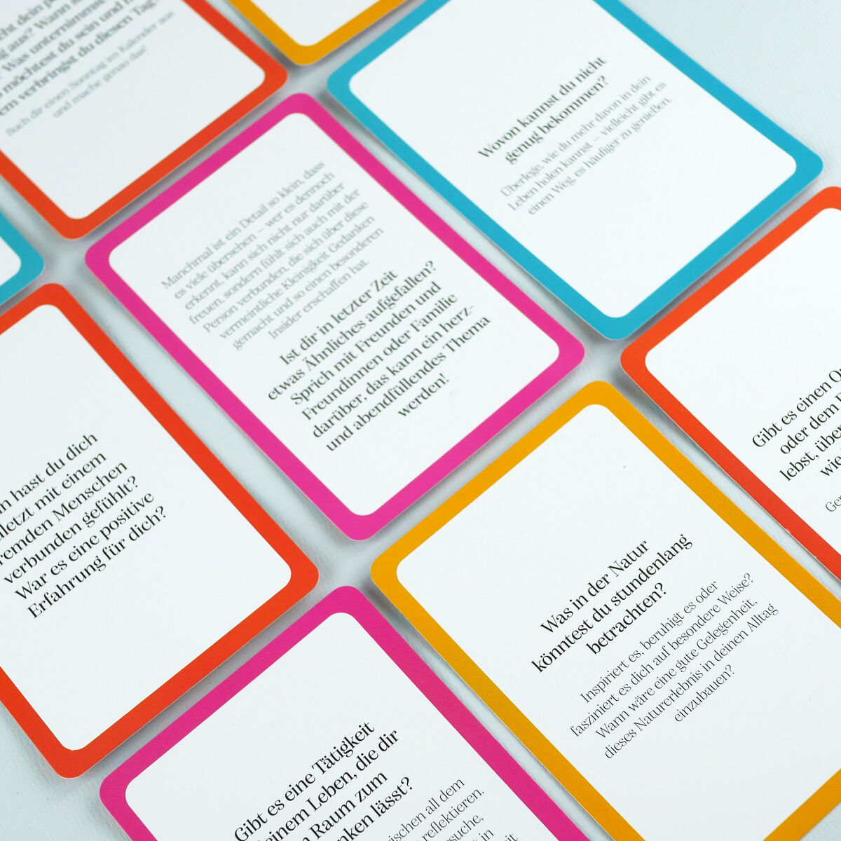 Mehrere Inspirations- und Reflexionskarten aus dem Set „Was mein Leben reicher macht“ von DIE ZEIT liegen versetzt auf weißem Hintergrund. Jede Karte zeigt einen bunten Rahmen in Gelb, Orange, Rosa oder Türkis und ist mit reflektierenden Fragen und Zitaten bedruckt.