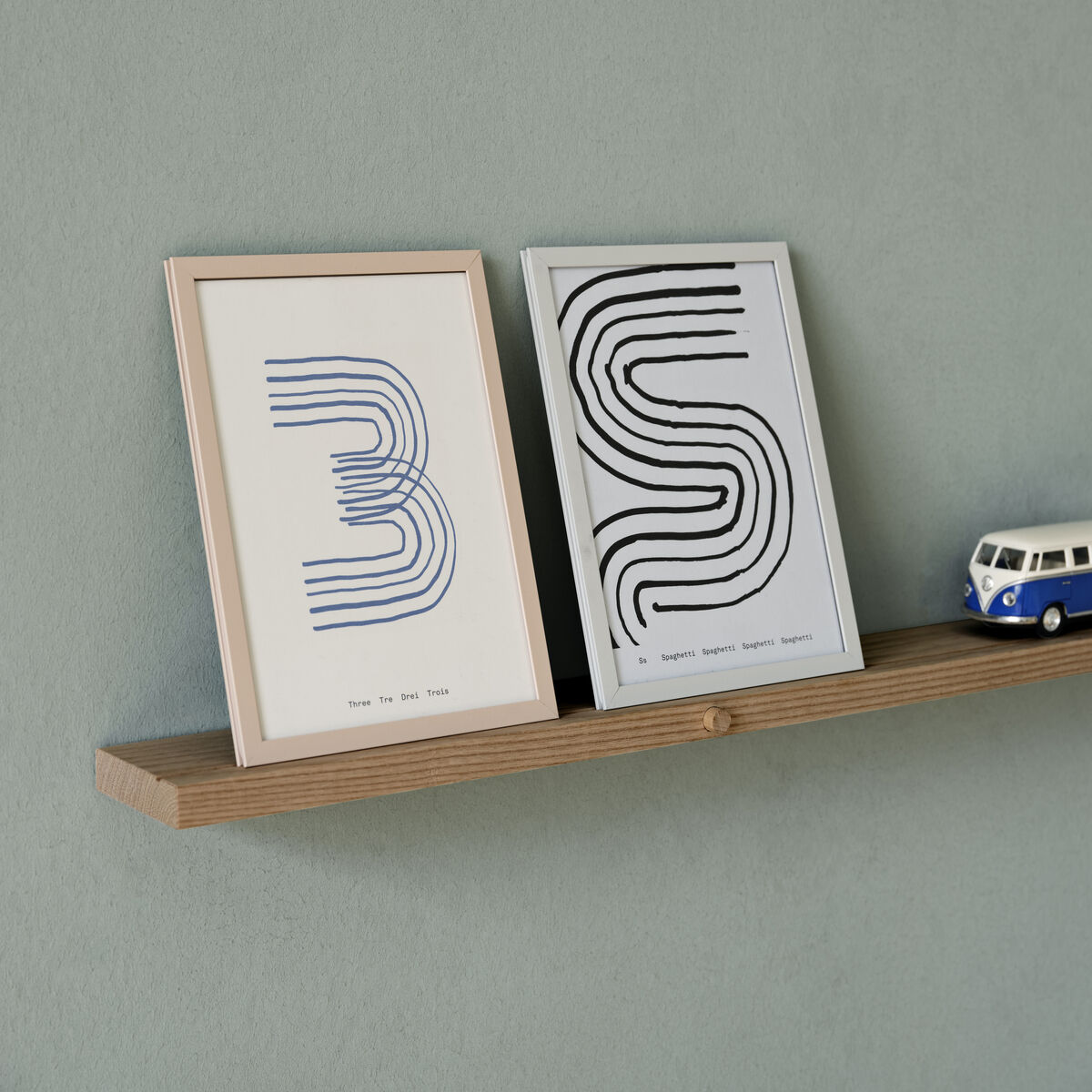 Ein minimalistisches Regal aus Eichenholz an einer Wand, auf dem zwei gerahmte Kunstdrucke mit abstrakten Linien und ein kleines blau-weißes Modellauto stehen.