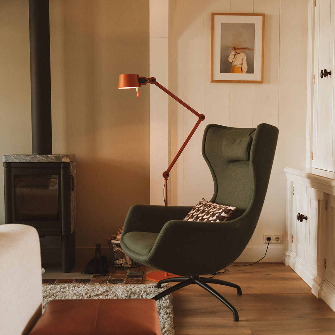 Modernes Wohnzimmer mit einer zweiarmigen Stehlampe »BOLD« in Orange, einem grünen Sessel, einem Kamin, und weißen Schränken. Die Lampe beleuchtet den Bereich neben dem Sessel, der ideal für Lese- oder Entspannungszeiten ist.