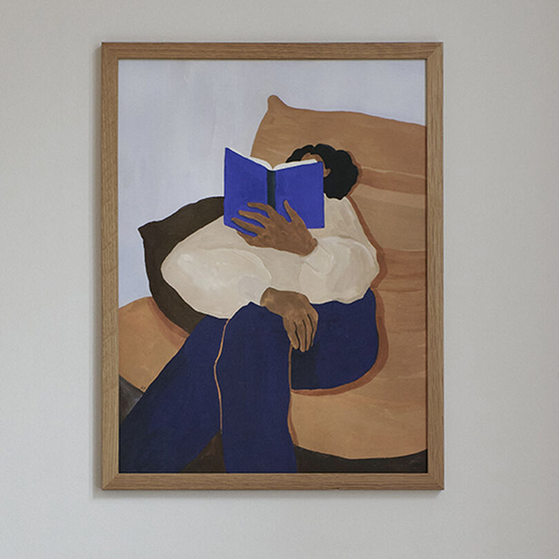 Ein Gemälde von Hanna Peterson mit dem Titel 'The Togo' in einem Rahmen. Das Bild zeigt eine lesende Person, die auf einem beigen Bett sitzt und ein blaues Buch hält. Im Vordergrund steht ein kleines rundes Tischchen mit einer grauen Kugel und einer Kegel-Dekoration.