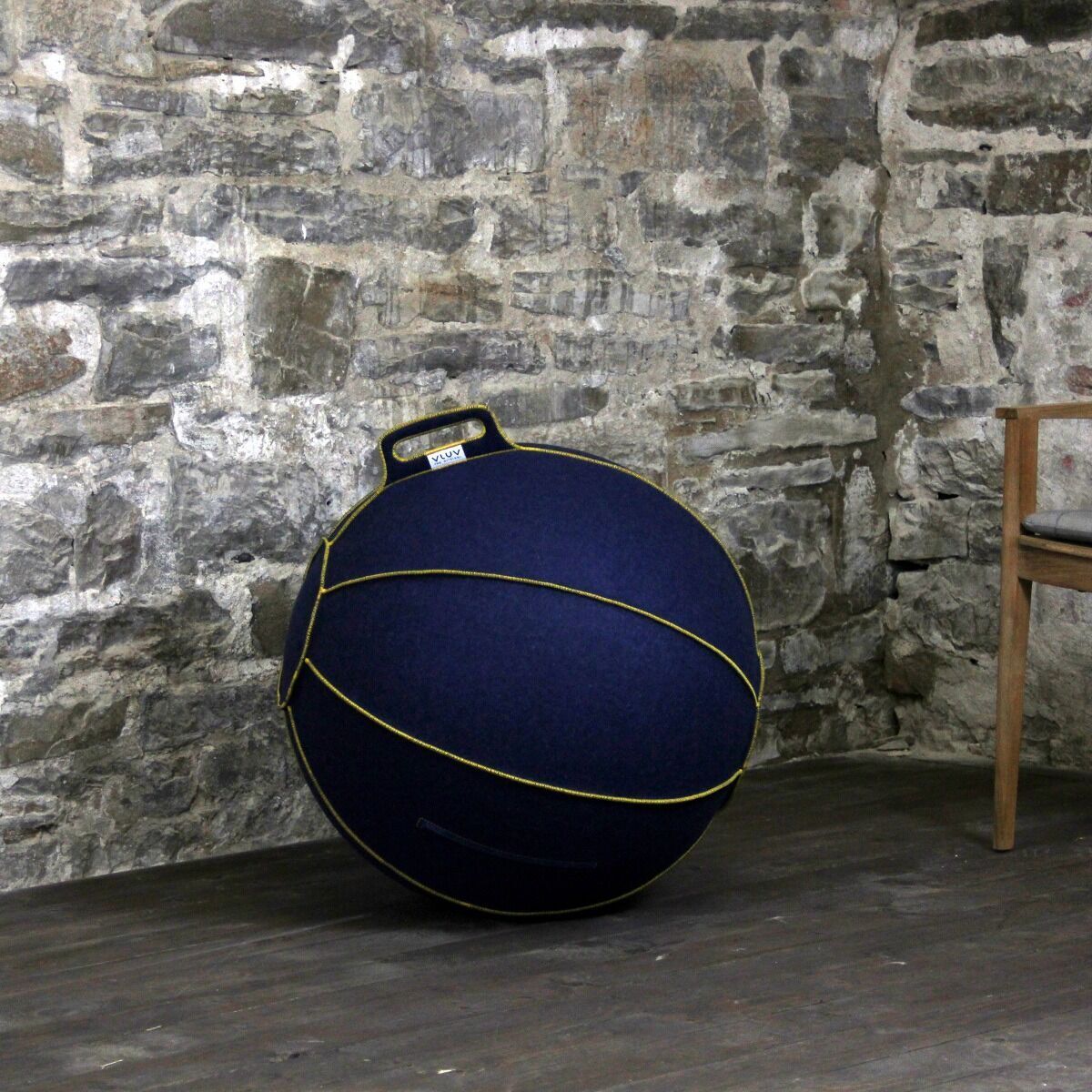 Ein grauer Sitzball (»Vluv Velt«) mit gelben Nähten und Griff, neben einem braunen Holzstuhl, vor einer rustikalen Steinwand.