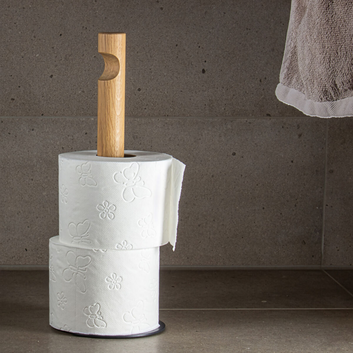 Der Küchenrollenhalter »GIVE« der Marke finbeaux steht auf grauem Fliesenboden. Er besteht aus einem runden schwarzen Sockel und einem senkrechten Griff aus hellem Holz mit charakteristischer Ausstanzung. Zwei Toilettenpapierrollen sind dekorativ gestapelt.
