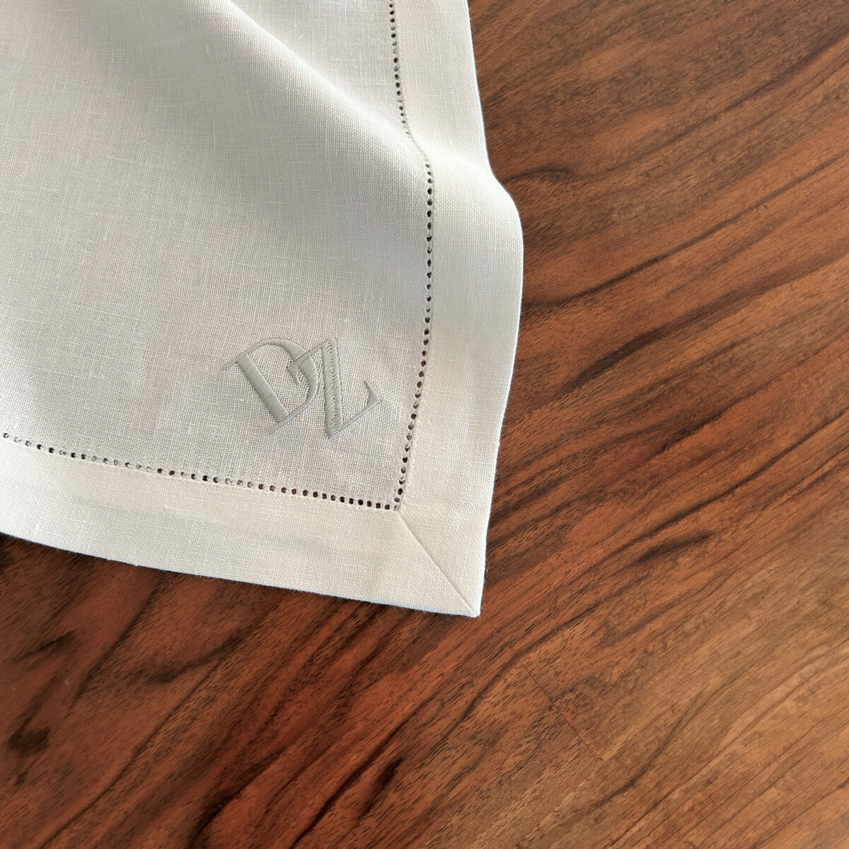 Eine weiße Leinenserviette liegt auf einer hölzernen Tischoberfläche. An einer Ecke der Serviette ist ein Monogramm mit den Initialen 'DZ' in das Gewebe gestickt. Die Serviette hat einen gestrickten Rand und wirkt hochwertig.