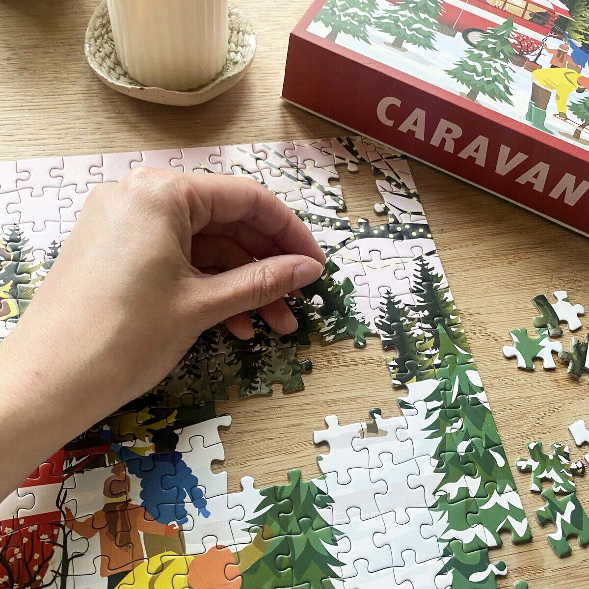 Eine Hand setzt ein farbenfrohes 300-Teile-Puzzle zusammen, das eine illustrativ gestaltete Szene mit Wohnwagen, Tannenbäumen und Menschen in bunten Outfits zeigt. Die Puzzle-Schachtel mit dem Titel „Caravan“ und eine angezündete Kerze stehen daneben auf einem Holztisch.