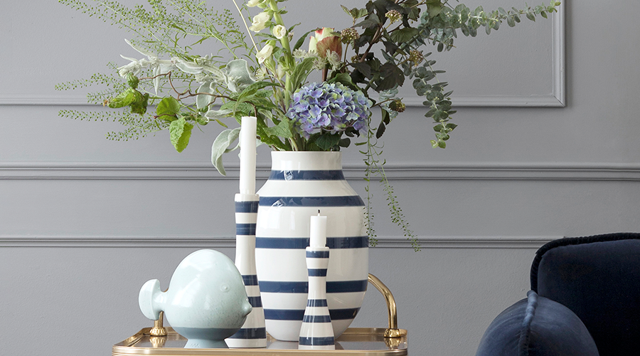 Vase und zwei Kerzenhalter mit blauen und weißen Streifen, gefüllt mit Blumen und grünen Zweigen, auf goldfarbenem Tablett vor grauer Wand