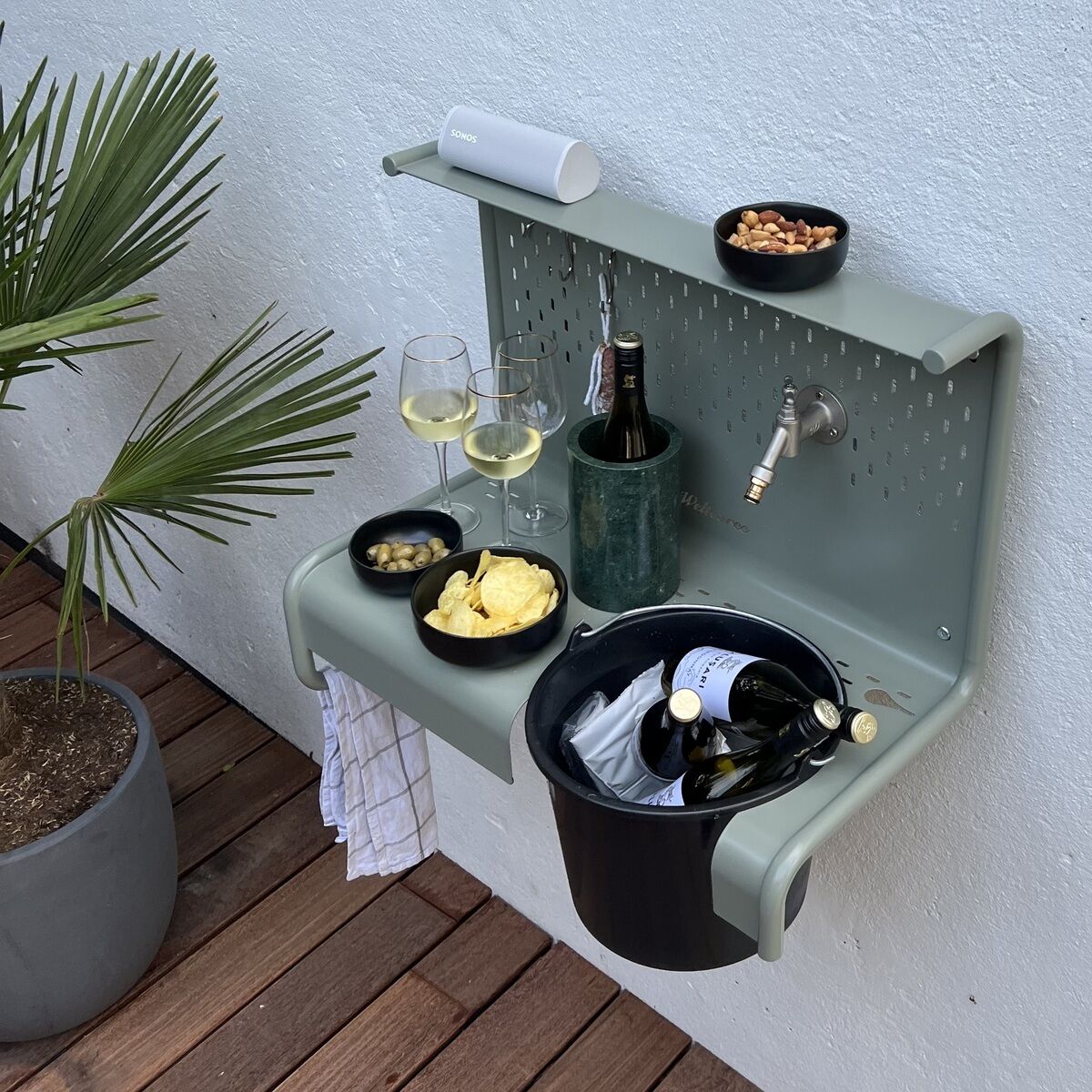 Die Gartenstation »Waterworks« von Weltevree ist an einer Außenwand montiert. Sie bietet einen integrierten Wasserhahn, eine Abstellfläche mit Getränkekühler, Gläsern, Snacks und Flaschen. Auf den Holzdielen steht eine Pflanze. Lichterketten sorgen für stimmungsvolle Beleuchtung.