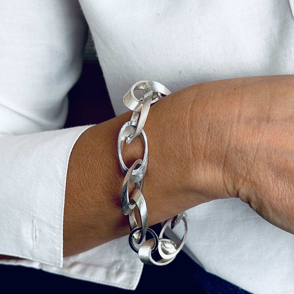 Nahaufnahme eines silbernen Kettenarmbands am Handgelenk einer Person, die ein weißes Hemd und eine blaue Hose trägt.