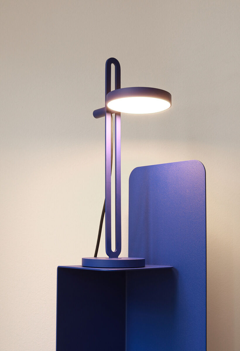 Foto einer Tischlampe namens »Echo«. Die Lampe hat einen minimalistischen blauen Metallrahmen und einen runden, weißen Lampenschirm. Sie steht auf einem blauen Möbelstück vor einer hellen Wand.