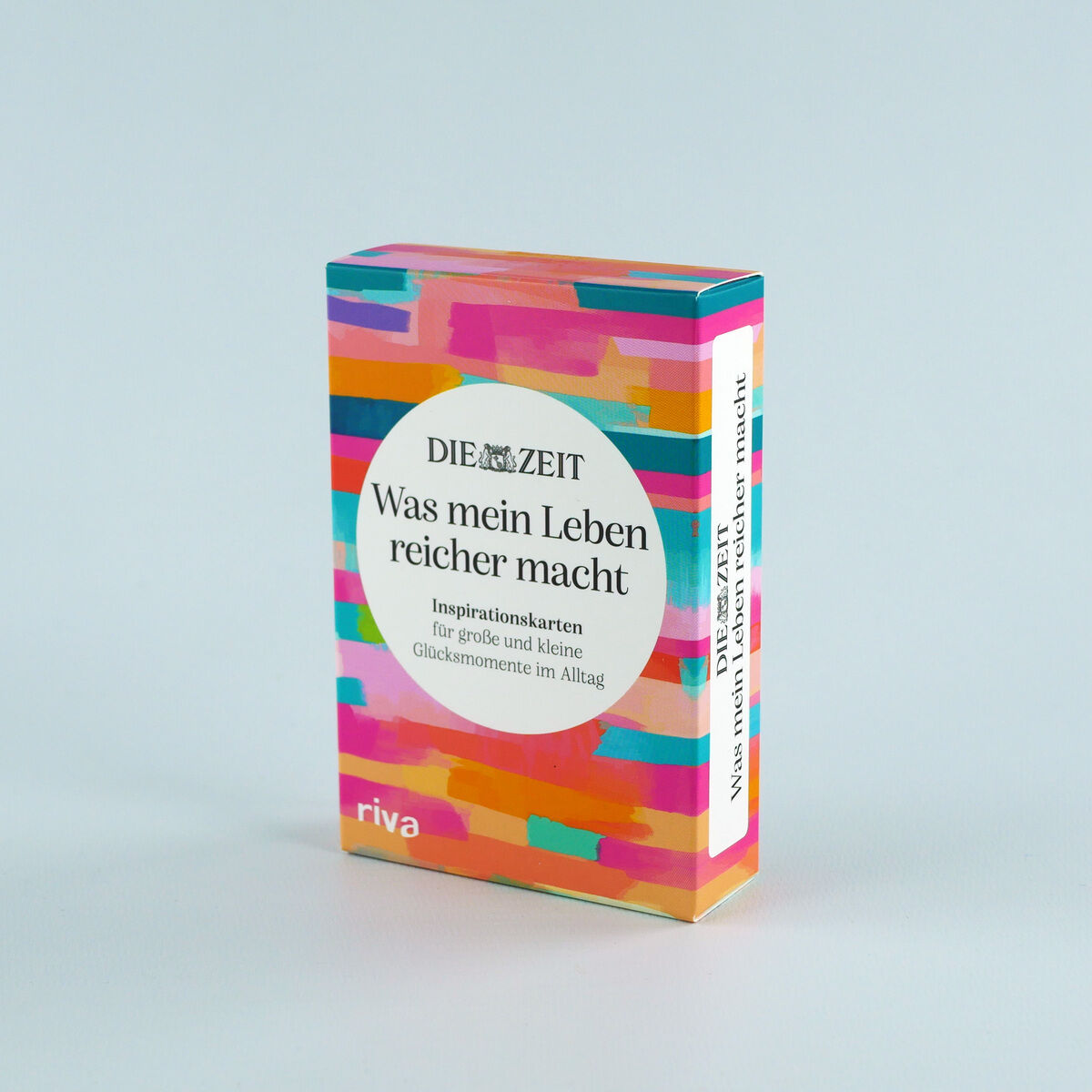 Bunte Box mit dem Titel »Was mein Leben reicher macht« und dem Logo von DIE ZEIT. Die Vorderseite zeigt einen weißen Kreis mit schwarzer Schrift, umgeben von lebhaft gemusterten Farbstreifen in Pink, Orange, Gelb, Türkis und Blau. Am unteren Rand steht „riva“.