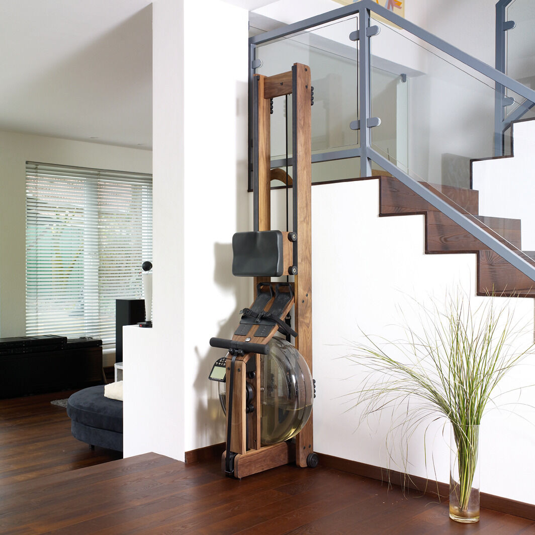 Ein Waterrower-Rudergerät aus Eiche steht an einer weißen Wand neben einer Treppe in einer modernen Wohnzimmereinrichtung. Das Gerät hat ein elegantes Holzdesign und passt farblich zur Umgebung. Eine Pflanze befindet sich auf dem Boden in der Nähe.