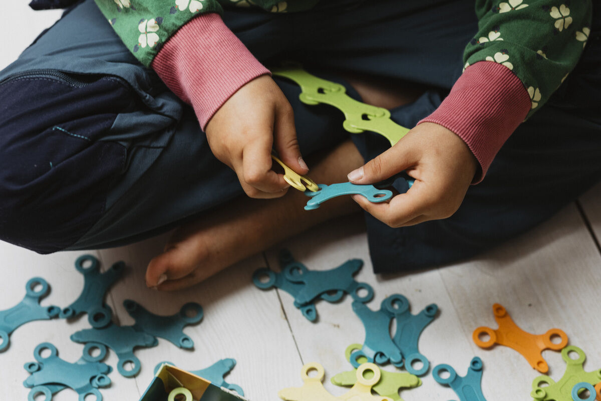 Ein Kind sitzt auf dem Boden und spielt mit farbigen Teilen des Spiels 'Binabo'. Die Teile sind hauptsächlich grün, blau und gelb.