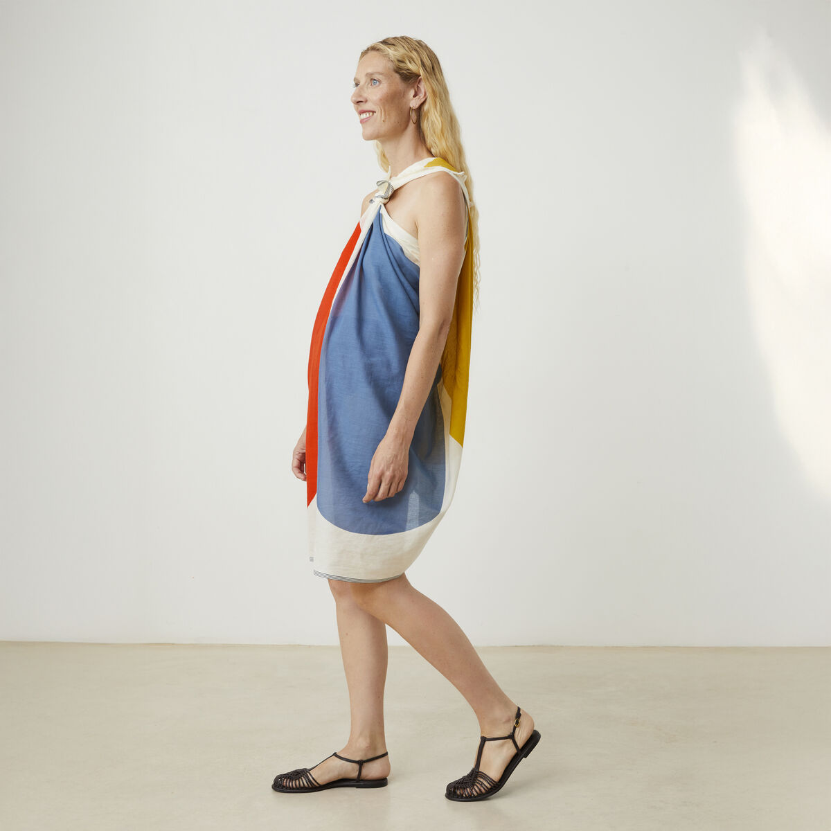 Frau mit dem Baumwollschal »No. 727« von Moismont: Der große Schal aus feiner Baumwolle zeigt ein auffälliges, grafisches Muster in Gelb, Rot, Rosa, Weiß und Blau. Sie trägt ihn als Kleid, geknotet an der Schulter, vor neutralem Hintergrund, barfuß in Sandalen.