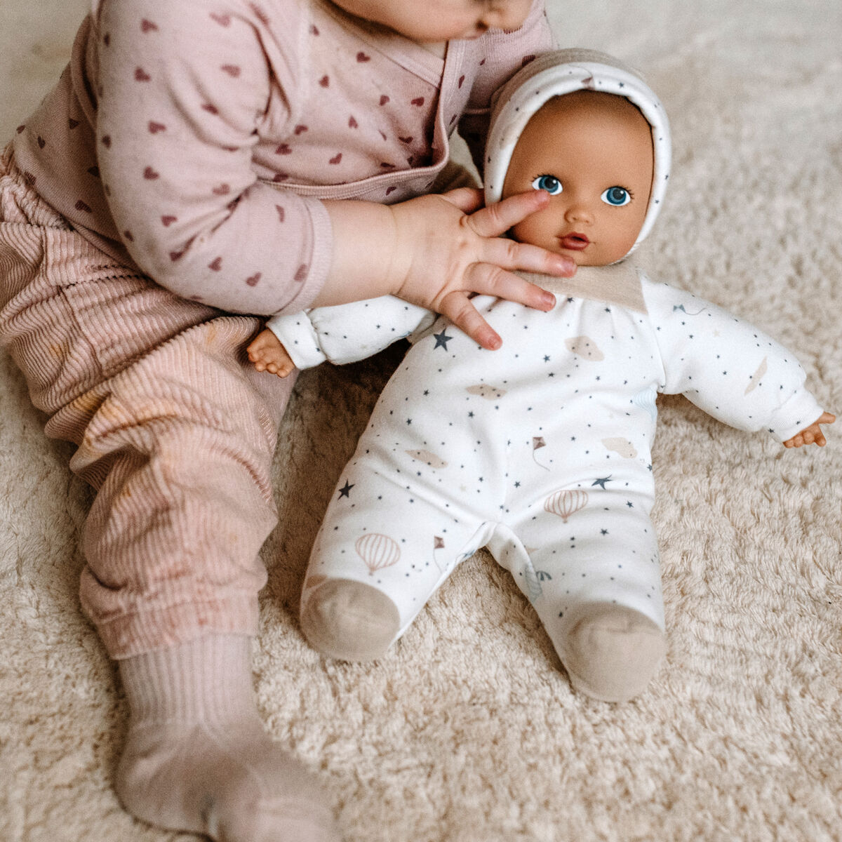 Ein Baby im rosafarbenen Schlafanzug sitzt auf einem cremefarbenen Teppich und hält eine Puppe im weißen Strampler. Die Szene wirkt ruhig, weich und gemütlich und zeigt ein kindgerechtes Geschenk aus der Geschenkbox »Baby Pure Sleep Well«.