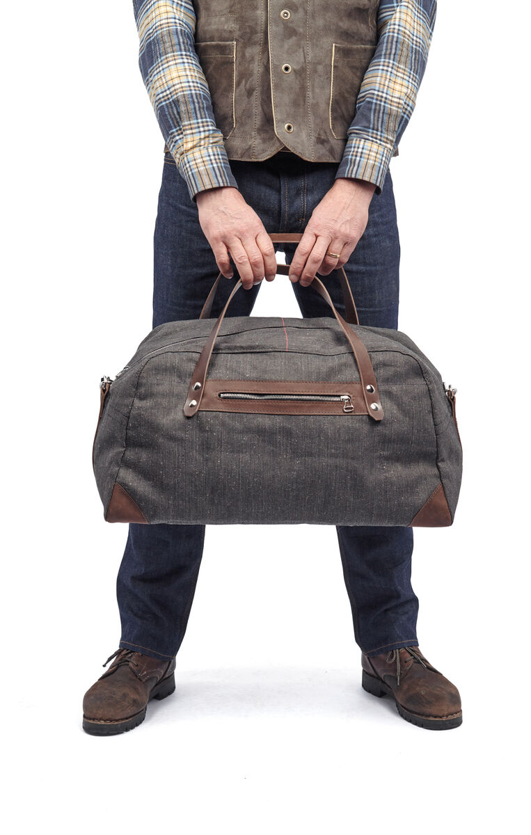 Eine männliche Person hält eine graue Weekender-Tasche aus Segeltuch mit braunen Lederdetails. Die Person trägt eine blaue Jeans, braune Schuhe, ein kariertes Hemd und eine braune Weste.