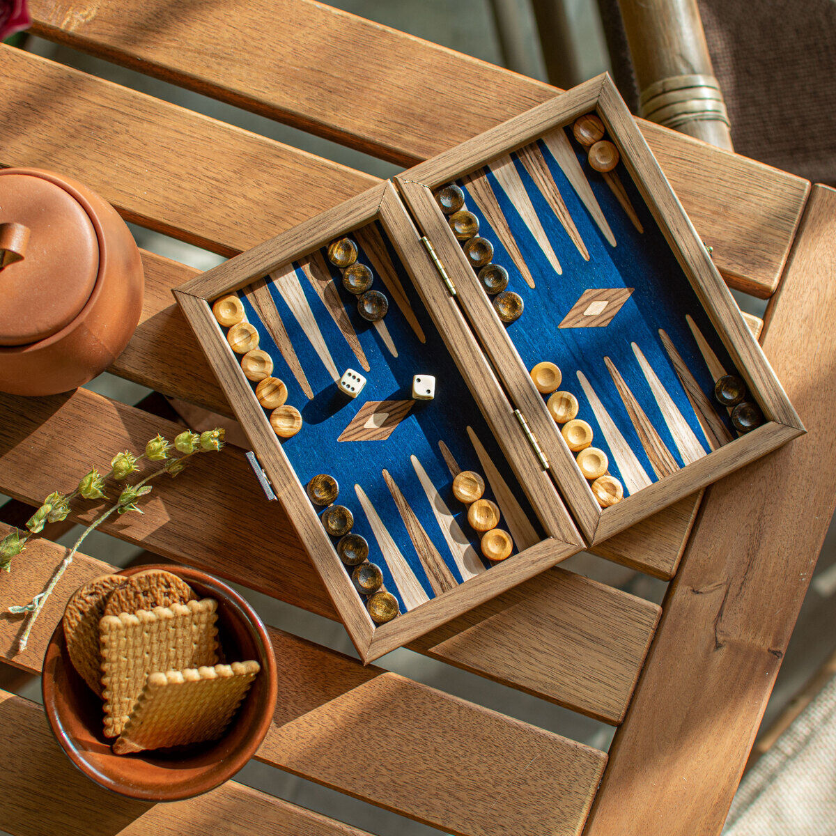 Ein Pocket-Backgammon-Brett aus Nussbaumholz liegt geöffnet auf einem Holztisch. Das Spielbrett hat ein blaues Spielfeld mit braunen und beige-gemusterten Steinen sowie zwei weißen Würfeln. Daneben stehen eine Teekanne, eine kleine Schale mit Keksen und ein kleiner Zweig als Dekoration.