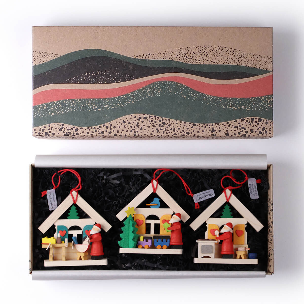 Eine Geschenkbox mit drei handgefertigten Holzweihnachtsmannhäusern als Baumschmuck. Jedes Haus hat kleine, farbenfrohe Details und hängt an roten Bändern. Die Box ist dekorativ gestaltet und enthält schwarze Füllung zum Schutz der Häuser.