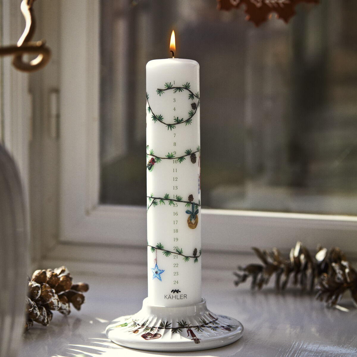 Die Adventskalenderkerze 2026 von Kähler steht auf einem dekorierten Teller am Fenster. Sie ist weiß mit grünem Tannengirlandenmuster, goldenen und roten Details sowie Adventskalenderzahlen. Daneben liegen Tannenzapfen und kleine rustikale Weihnachtsdeko.