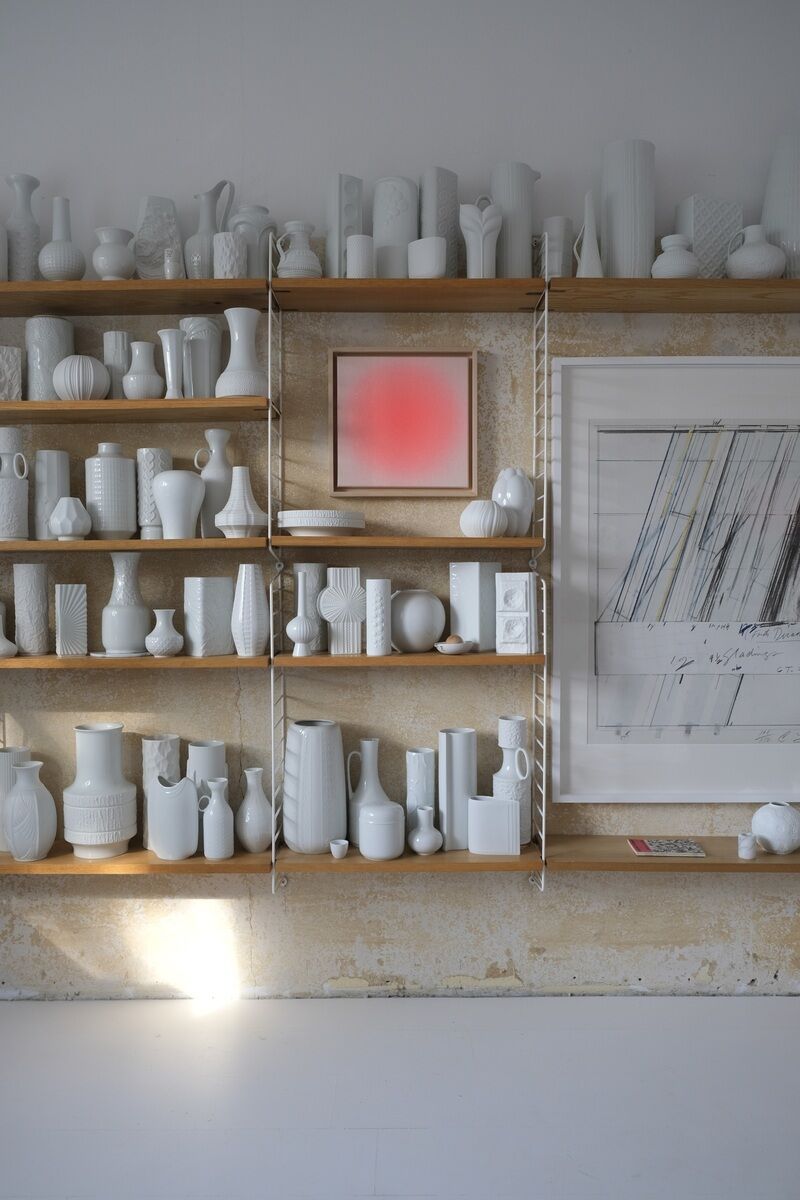 Ein heller Raum mit weißen Wänden und weißen Holzregalen, die mit zahlreichen weißen Keramikvasen und Skulpturen bestückt sind. Im Zentrum hängt ein quadratisches Kunstwerk mit einem roten Sorbet-Farbverlauf. Rechts daneben ist eine große Grafik zu sehen. Der Boden ist weiß und ein wenig Tageslicht scheint von links in den Raum.