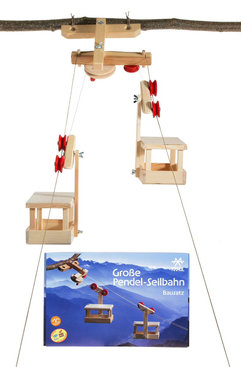 Große Pendelseilbahn Bausatz mit zwei Wagen und Bergstation aus Holz. Das Spielzeug ist an einem Ast befestigt. Im Hintergrund ist die Verpackung mit einer blauen Berglandschaft abgebildet. Das Set fördert Kreativität und motorische Fähigkeiten.