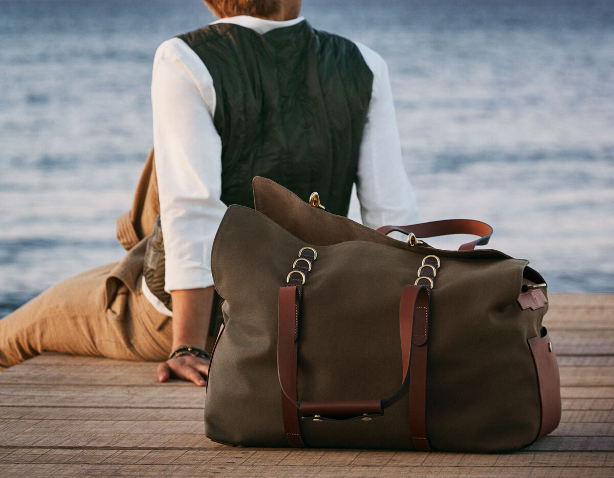 Eine große braune Reisetasche mit Ledergriffen steht auf einem Holzsteg am See. Im Hintergrund sitzt eine Person und schaut übers Wasser. Die Farben der Tasche und der Umgebung sind natürlich gehalten.