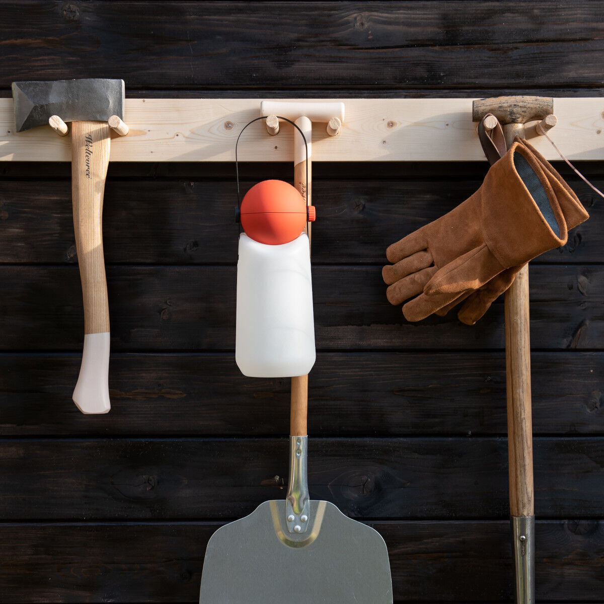 Die »Guidelight« Lampe hängt an einer Holzlatte, umgeben von verschiedenen Werkzeugen wie einer Axt, einem Paar Arbeitshandschuhe und einer Schaufel. Die Lampe hat einen weißen Körper und einen orangen Griff.