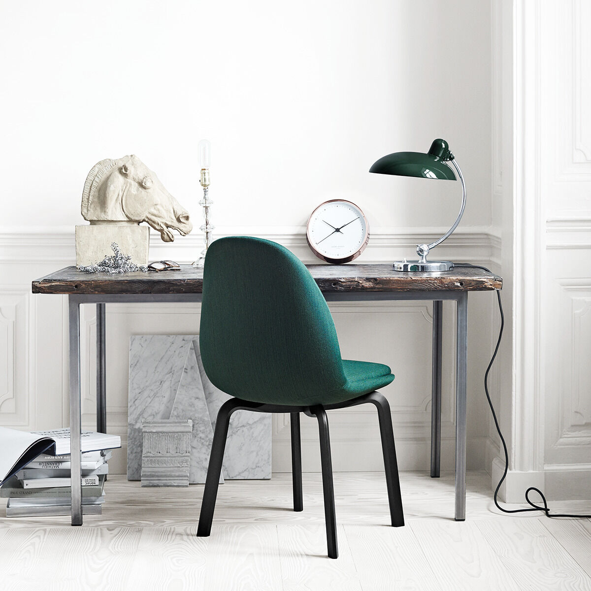 Ein grüner Stuhl und eine Schreibtischlampe »Kaiser Idell« stehen auf einem Schreibtisch in einem minimalistischen, hellen Raum mit weißen Wänden und Holzboden. Auf dem Schreibtisch befinden sich Dekorationen und eine Uhr.
