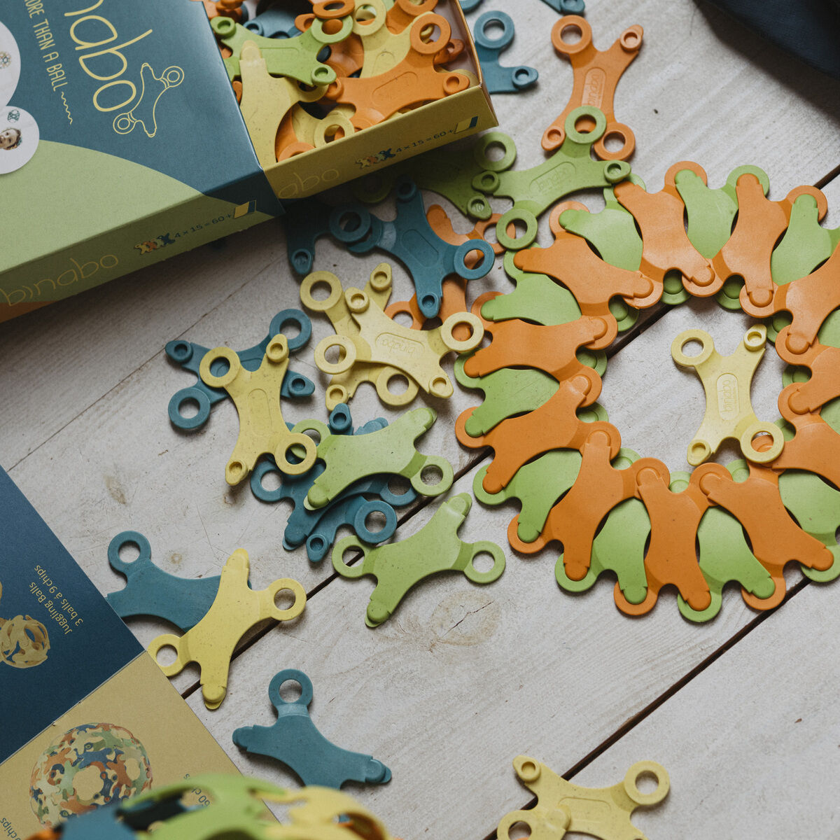 Das Bild zeigt das Spiel »Binabo« mit 60 Teilen. Verschiedene grüne, gelbe, orange und blaue Steckteile wurden teilweise miteinander verbunden und liegen ausgebreitet auf einem hellen Holzboden. Die Verpackung des Spiels ist ebenfalls sichtbar.