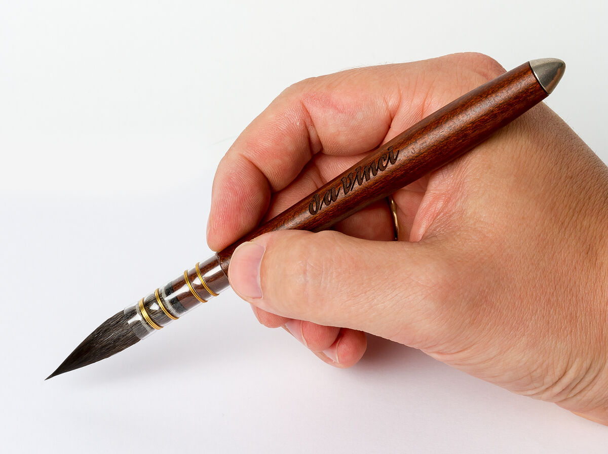 Ein traditioneller Aquarellpinsel mit Ethergraf®-Metallspitze, gehalten von einer Hand. Der Pinsel hat einen Holzgriff und Metallakzente. Geeignet für feine Detailarbeit und Präzision in der Kunst.