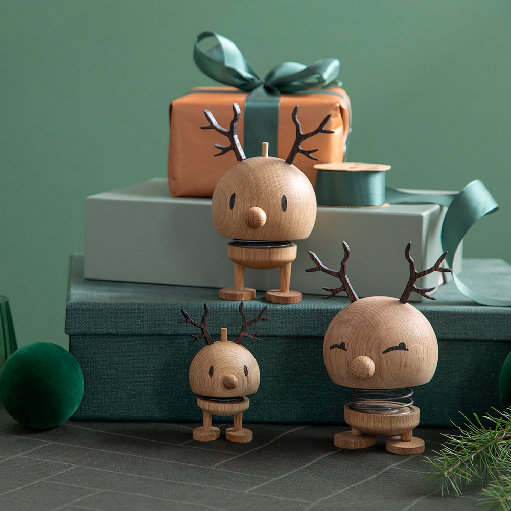 Drei hölzerne Rentierfiguren mit Federn als Beine und Geweihen, davor grüne Geschenkboxen und Dekoration.