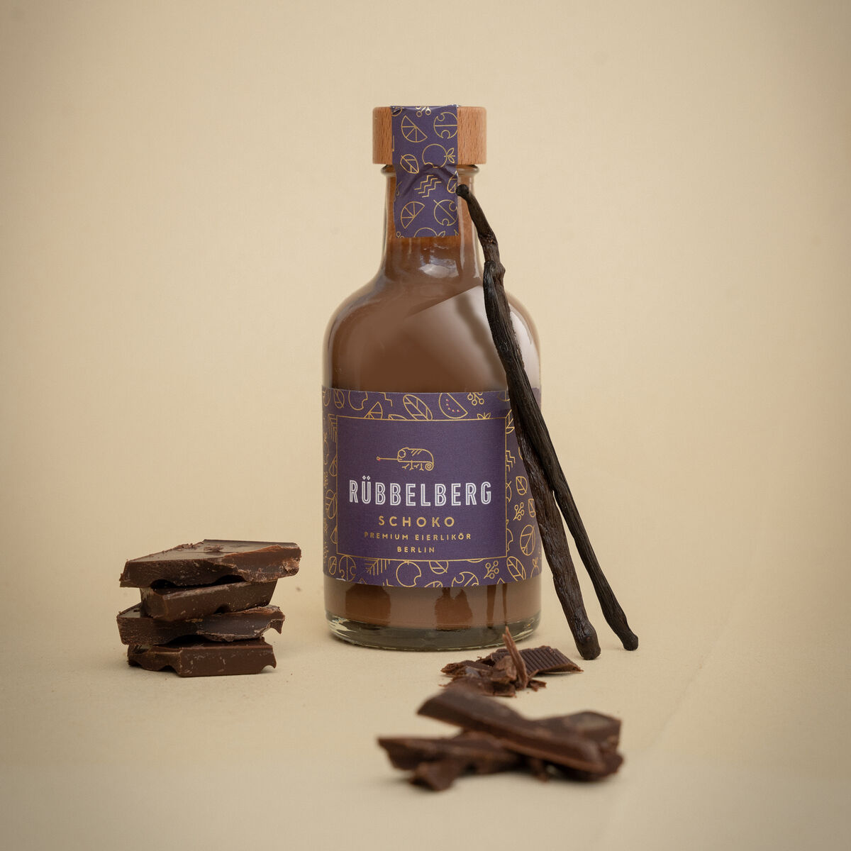 Flasche Premium Eierlikör Schoko der Marke Rubbelberg mit violettem Etikett, daneben Vanilleschoten und gestapelte Schokoladenstücke auf beigem Hintergrund.