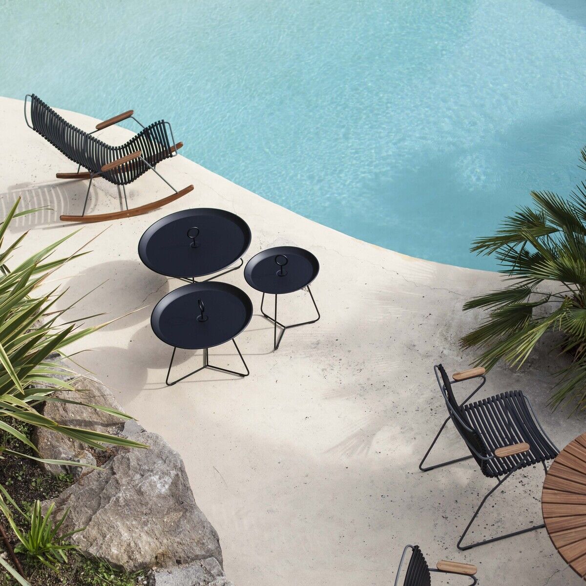 Eine entspannte Szene im Freien mit einem modernen schwarzen Schaukelstuhl, einem Pool und mehreren kleinen runden Tischen sowie bequemen Sitzgelegenheiten auf einer hellen Terrasse, umgeben von grünen Pflanzen und Palmen.