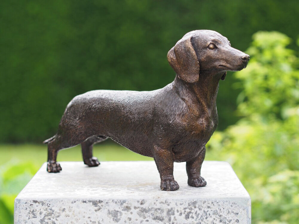 Bronzeskulptur eines Dackels auf einem grauen Steinpodest, umgeben von grünem Hintergrund in einem Garten.