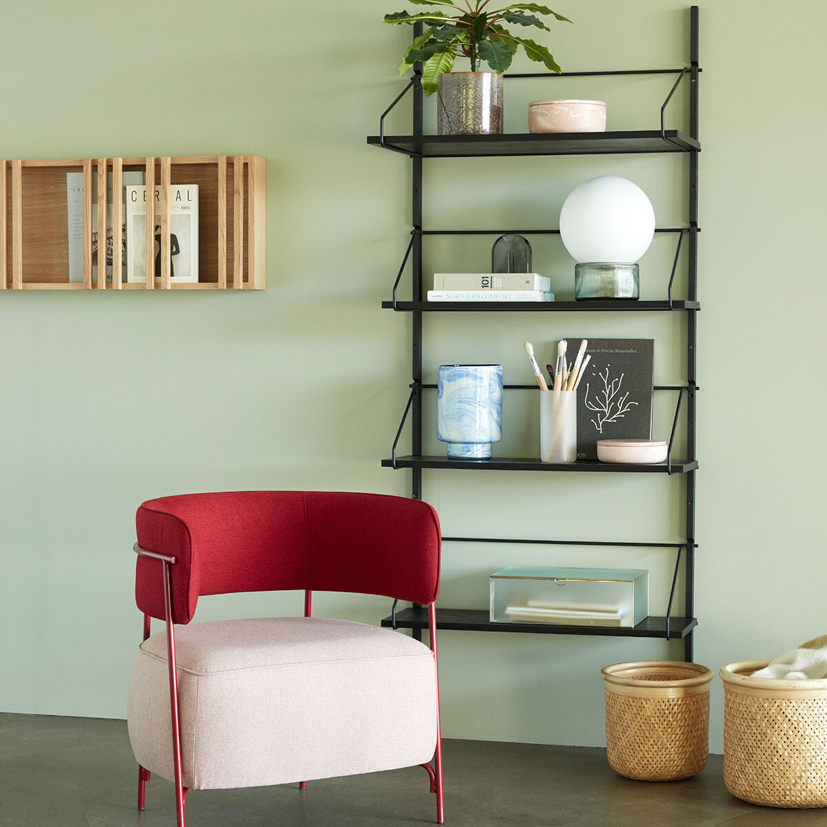 Ein modernes Wohnzimmer mit einem Magazinhalter aus Eichenholz an der Wand, neben einem schwarzen Regalsystem mit Büchern und Dekorationen. Eine rote und beige stoffbezogene Sessel steht vor dem Regal. Zwei Korbbehälter stehen auf dem Boden.