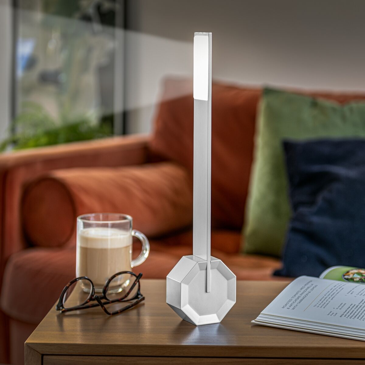 Eine oktagonförmige Tischlampe mit dem Namen 'One' steht auf einem Holztisch. Im Hintergrund sind ein Sofa mit roten und grünen Kissen sowie eine Brille und eine heiße Getränketasse zu sehen. Die Lampe wirkt modern und minimalistisch und scheint ein dimmbares LED-Licht zu besitzen.