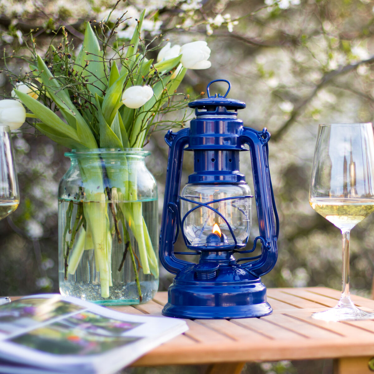 Eine blaue Sturmlaterne aus Stahl auf einem Holztisch im Freien, neben einem Glas Wein und einem Blumenstrauß in einer Vase.