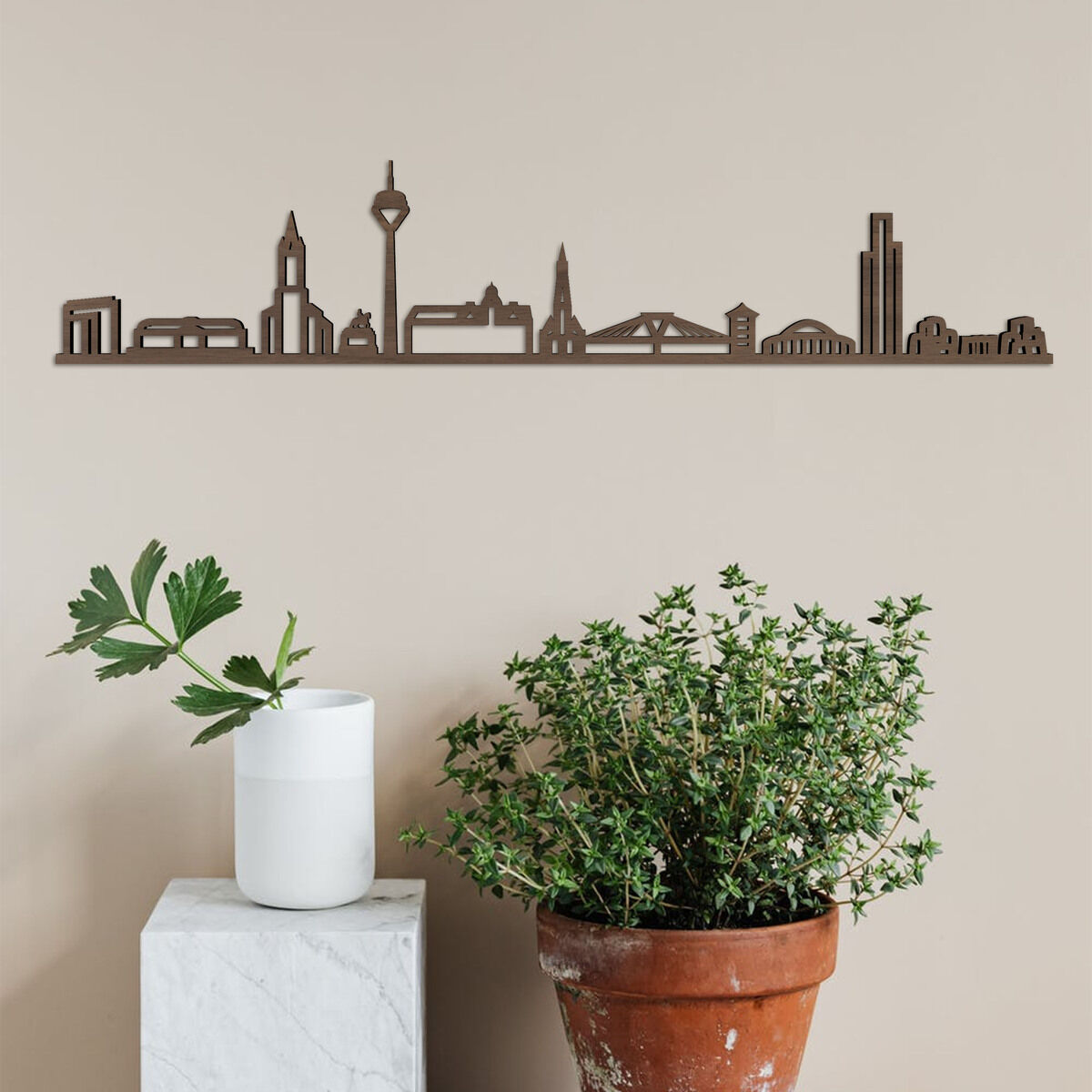 Eine hölzerne Skyline von Düsseldorf an einer beigen Wand montiert. Darunter ein hoher Blumentopf mit einer grünen Pflanze und ein kleinerer, weißer Topf mit einer weiteren Pflanze.