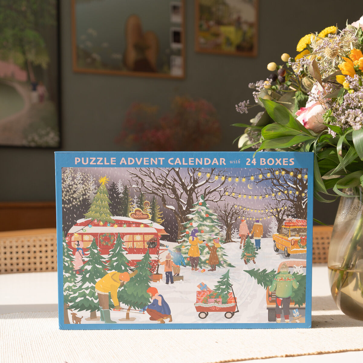 Eine bunt illustrierte Box mit dem Titel 'Puzzle Adventskalender mit 24 Boxen'. Das Design zeigt eine winterliche Szene mit festlich geschmückten Tannenbäumen, Menschen beim Kauf und Schmücken von Weihnachtsbäumen, beleuchtete Girlanden und Schneefall. Die Verpackung steht auf einem hellen Untergrund neben einem Glasvase mit Blumen. Das Bild vermittelt eine gemütliche, vorweihnachtliche Stimmung.