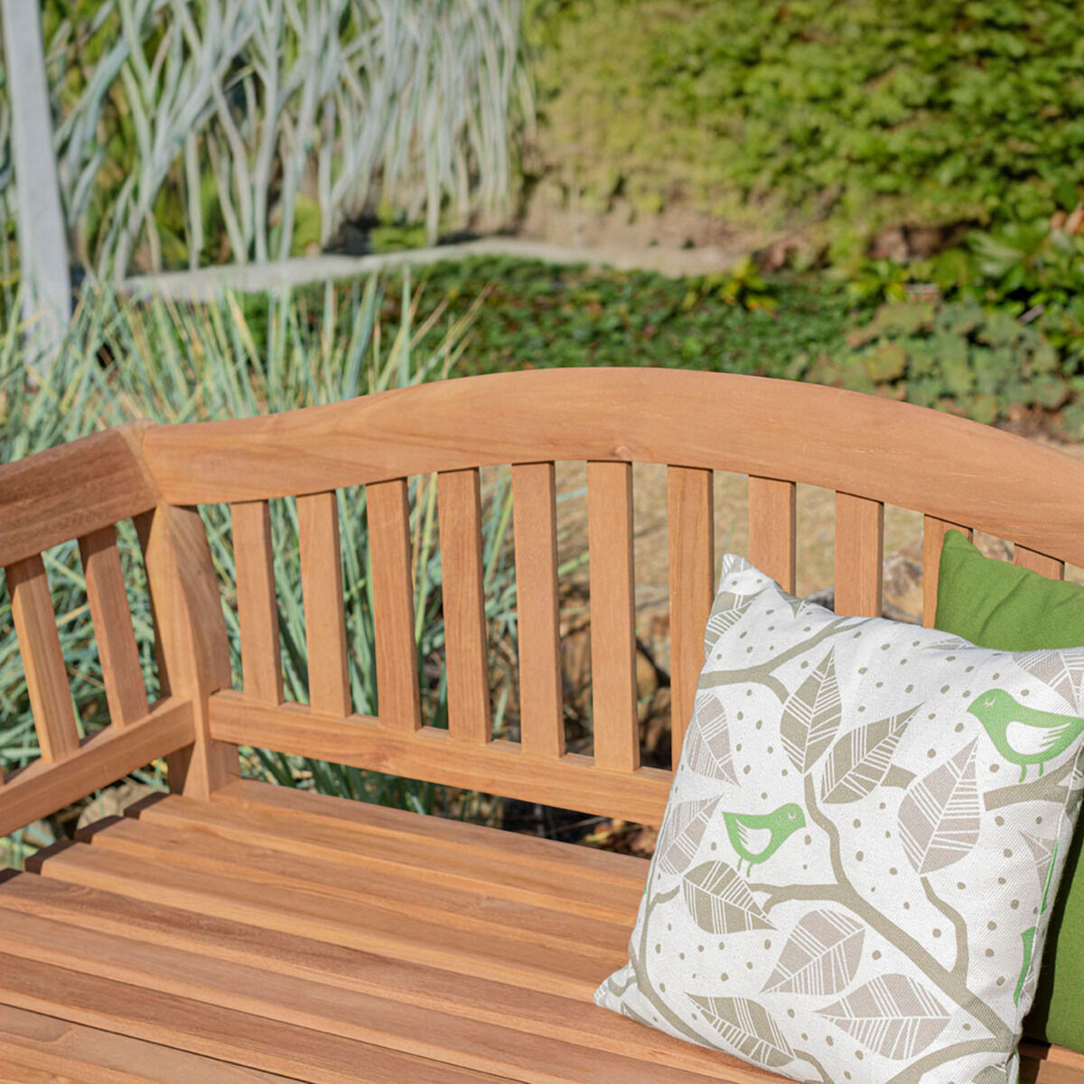 Nahaufnahme der Teak-Friesenbank »Wave« von bene living im Garten. Die unbehandelte, warmbraune Holzbank zeigt eine sanft geschwungene Rückenlehne, senkrechte Streben und breite Sitzlatten. Rechts liegen ein weiß-grau gemustertes Kissen mit grünen Tiermotiven und ein grünes Kissen; dahinter wachsen Gräser und Bodendecker.