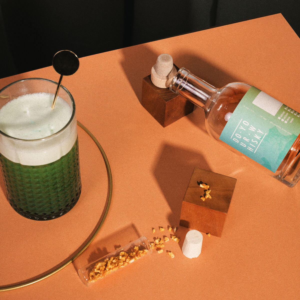 Ein Whisky-Set zum selber herstellen. Das Bild zeigt ein Glas mit grünem Getränk, einen Rührstab, eine Flasche mit der Aufschrift 'DIY to DRINK WHISKY', Holzstücke und ein Korken auf einem orangen Untergrund.