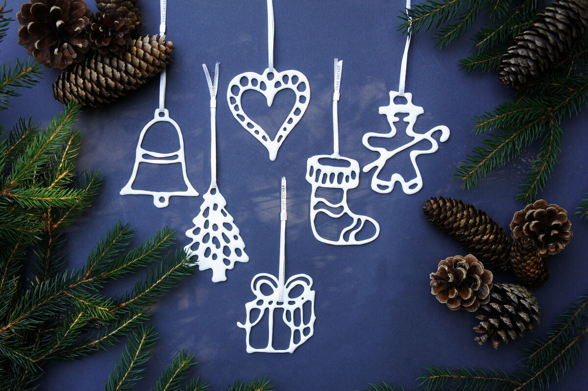 Ein Set von sechs Weihnachtsanhängern in verschiedenen Formen (Glocke, Herz, Weihnachtsbaum, Strumpf, Geschenk, Lebkuchenmann) hängt an einer Schnur, umgeben von Tannenzweigen und Tannenzapfen auf einem dunkelblauen Hintergrund.