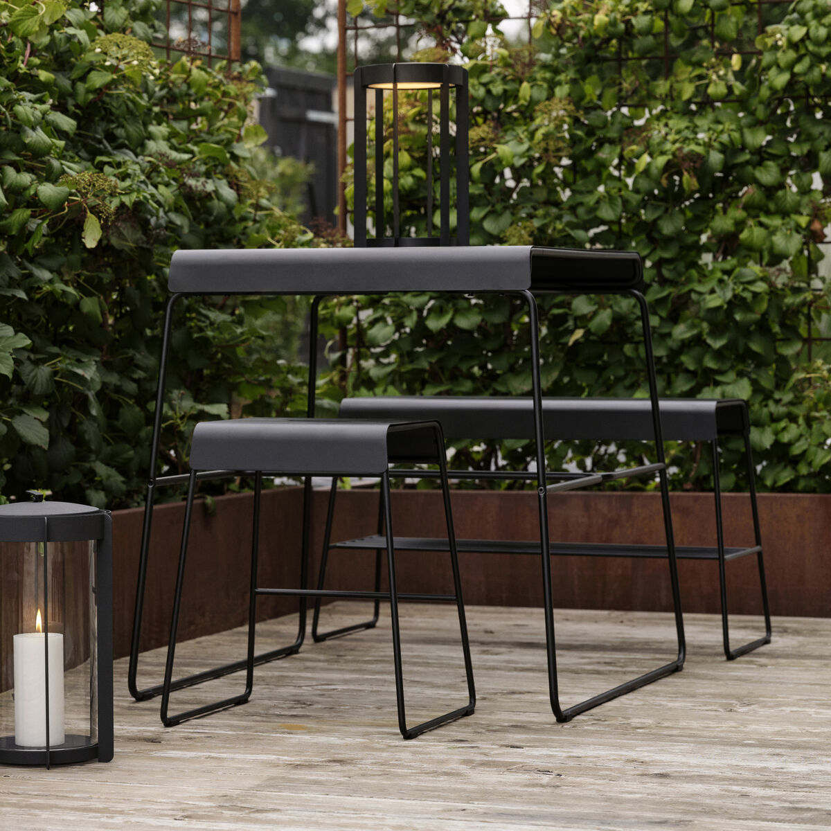 Moderne Outdoor-Bank mit Ablage auf einem Holzdach, umgeben von grüner Vegetation und zwei Laternen mit Kerzen, die eine gemütliche Atmosphäre schaffen.