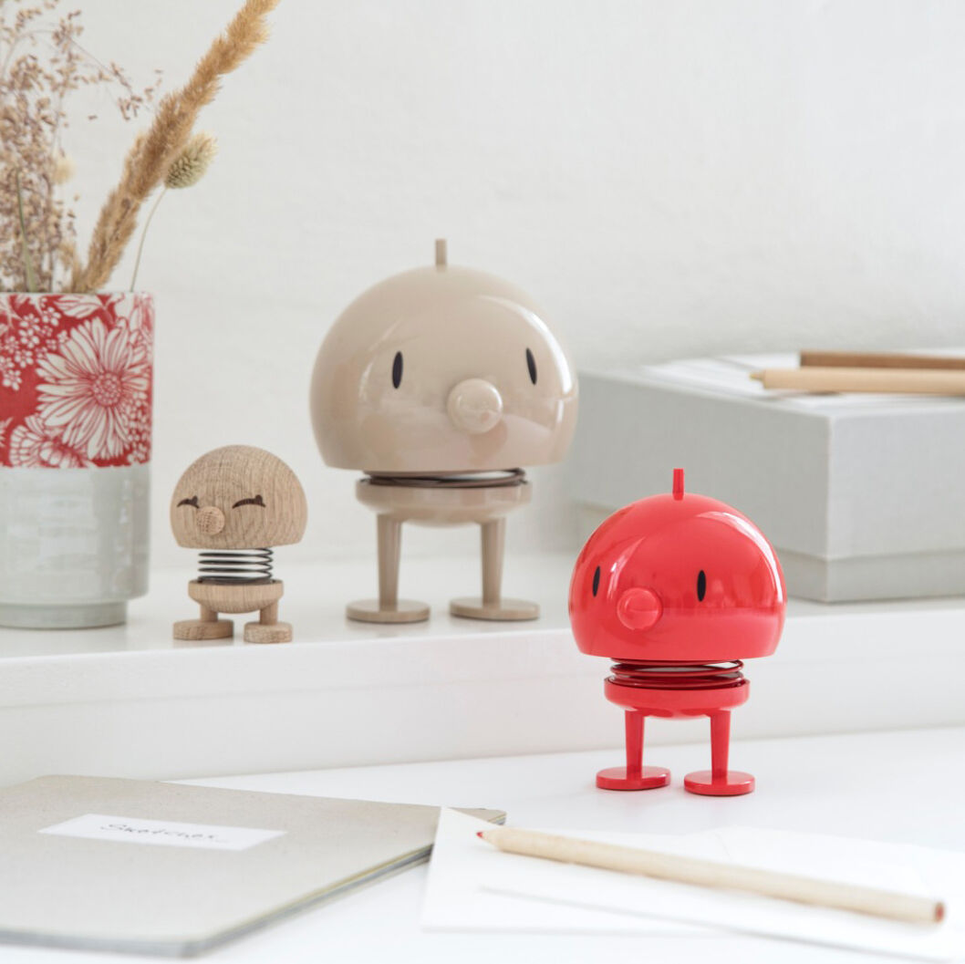 Bunte Hoptimist Bumble Figuren, darunter eine rote, stehen auf einem weißen Regal. Dahinter befinden sich ein beige Hoptimist und kleine dekorative Gegenstände wie Blumen in einer Vase und Stifte.