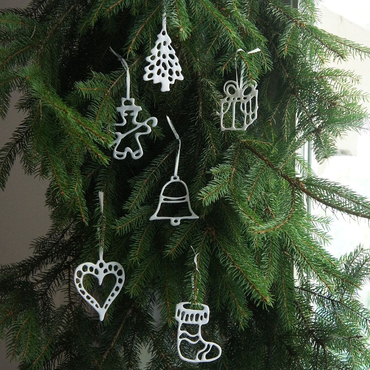 Sechs feine, weiße Weihnachtsanhänger aus Bonechina-Porzellan in Form von Herz, Tanne, Lebkuchenmann, Geschenkpaket, Glocke und Stiefel hängen an den Ästen eines grünen Nadelbaums vor einem hellen Fenster. Die filigranen Motive wirken elegant und winterlich.