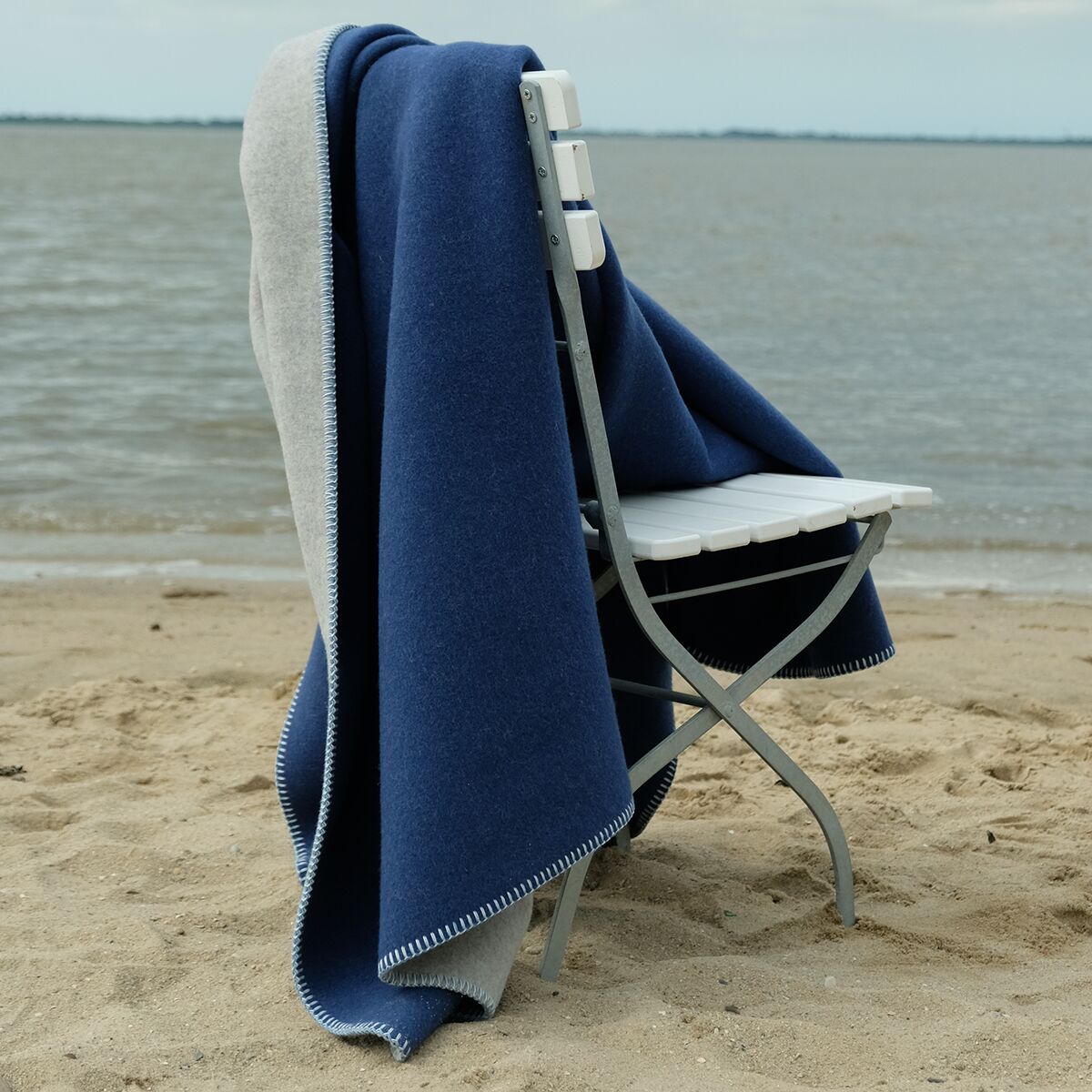 Eine zweifarbige Merinowolldecke in Blau und Grau liegt locker auf einem weißen Stuhl am sandigen Strand mit Blick aufs Meer. Die Szene vermittelt eine ruhige, gemütliche Atmosphäre am Wasser und betont die weiche, hochwertige Textur der Decke.