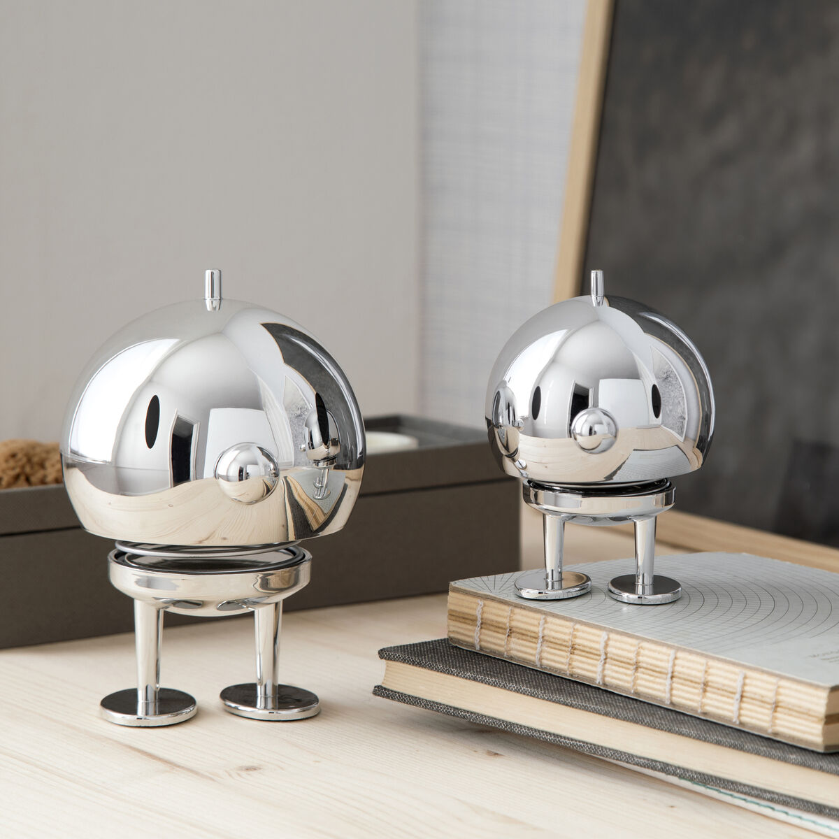 Zwei glänzende Hoptimist Bumble Chrome Figuren aus Metall stehen auf einem Tisch. Eine größere und eine kleinere Figur, beide mit abgerundeten Formen und minimalistischen Design. Im Hintergrund sind ein paar Bücher und dekorative Elemente sichtbar.