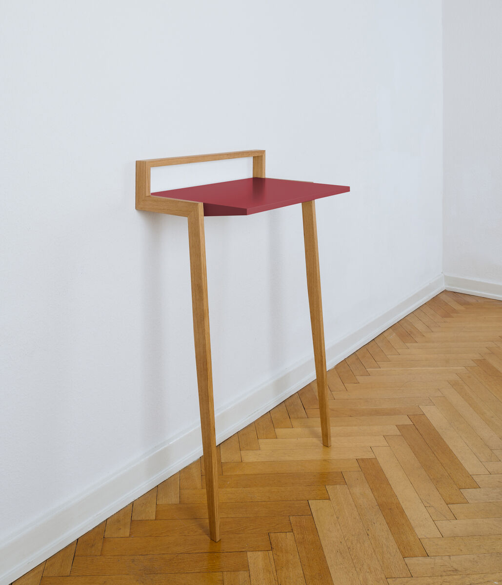 Ein modernes Stehpult mit einer roten Arbeitsfläche und natürlichen Holzbeinen, das für die Wandmontage konzipiert ist. Es steht auf einem hölzernen Parkettboden vor einer weißen Wand.